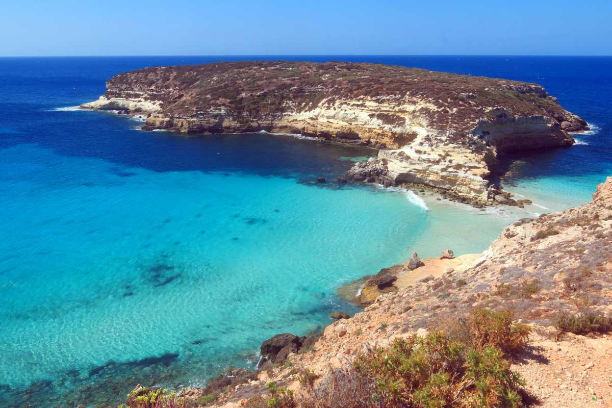 Vista panoramica di Cala Guitgia all'alba, con sabbia dorata, mare turchese e barche di pescatori. In lontananza si intravedono alcune strutture alberghiere e il profilo del centro abitato di Lampedusa.